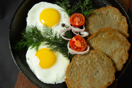 hot fried eggs with potato pancakes on a cast iron frying pan together with greens and vegetables on a black backgroundの写真素材