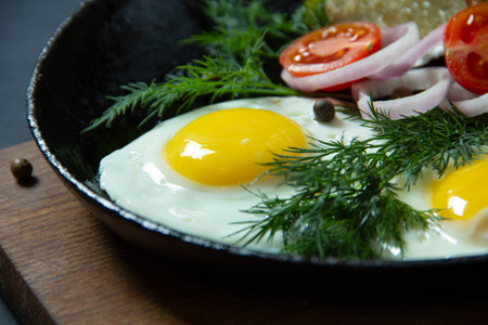 fried eggs with potato pancakes on a cast iron frying pan together with greens and vegetables on a black backgroundの写真素材