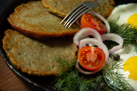 fried eggs with potato pancakes on a cast iron frying pan together with greens and vegetables on a black backgroundの写真素材