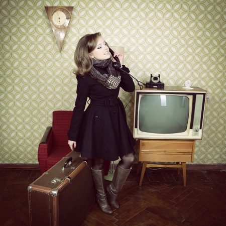 art portrait of young woman standing in room calling phone with vintage wallpaper and interior with tv, clocks, chair and suitcase, retro stylization 60-70s, tonedの写真素材