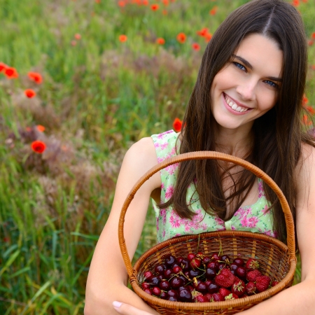 Young beautiful woman holding basket with cherry and strawberry on a poppy field, summer nature outdoor.の写真素材