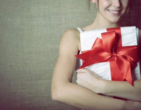 Portrait of attractive cheerful girl in sleeveless sports white shirt holding gift box with red bow over canvas background, toned and noise addedの写真素材