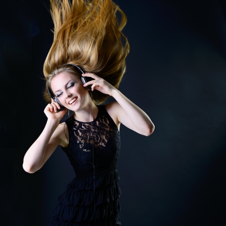 Music girl. Young beautiful excited woman with headphones listening music and dancing with long blond hair fly-away against dark background の写真素材