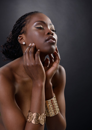 Beautiful young african woman posing at studio in golden jewellery, face with hand portrait over dark backgroundの写真素材
