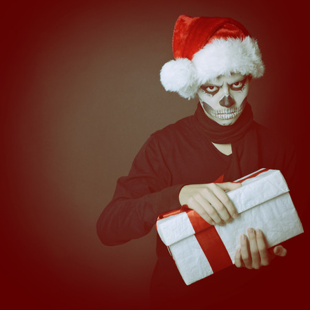 Holiday background of halloween person with terrible skull make-up in santa's hat opening gift box, tonedの写真素材