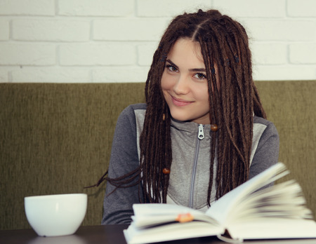 Young woman drinking coffee and reading book sitting indoor in urban cafe. Cafe city lifestyle. Casual portrait of teenager girl. Toned.の写真素材