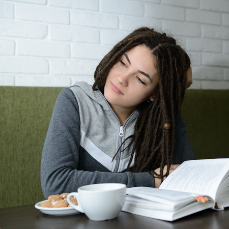 Young woman drinking coffee and reading book sitting indoor in urban cafe. Cafe city lifestyle. Casual portrait of teenager girl.の写真素材