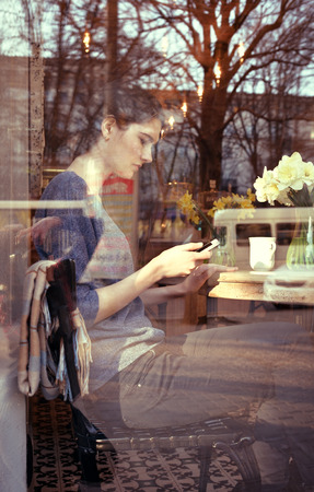 Young woman drinking coffee and use her smartphone sitting indoor in urban cafe. Cafe city lifestyle. Casual portrait of beautiful girl with spring flowers. Image toned.の写真素材