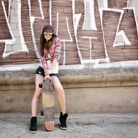 Portrait of beautiful teen girl standing on skateboard over wall with abstract graffiti art. Urban outdoors, teenager's lifestyle. Toned.の写真素材