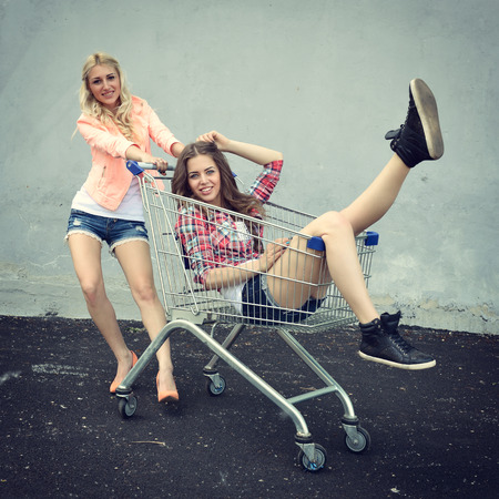 Two happy beautiful teen girls driving shopping cart outdoor, Image toned and noise added.の写真素材