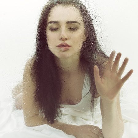 Beautiful young sensual woman is in bed on white sheet behind wet window, image toned.の写真素材