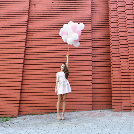 happy young woman standing over red brick wall and holding pink and white balloonsの写真素材