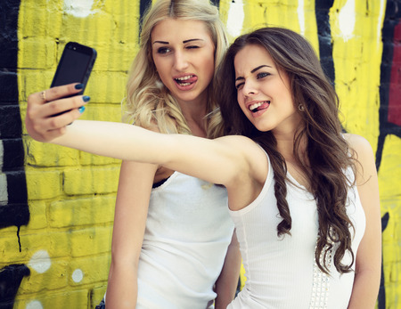 Happy attractive girls with smart phone take selfie against urban grunge graffiti wall.の写真素材