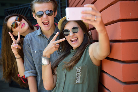 Young girls and boy having fun outdoor and making selfie with smart phone against red brick wall....