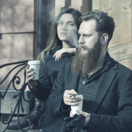 Autumn outdoor fashion portrait of young couple drinking coffee and smoking outdoor, image toned and...