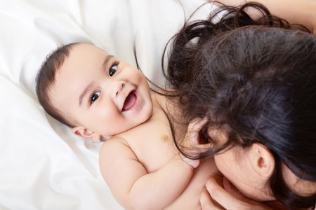 Young mother playing with her baby on white silk bed sheet. Cute mixed race bab boy has fun with mum, high key.の写真素材