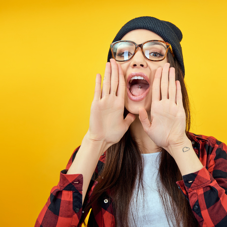 Hipster girl screaming like in megaphone holding hands near her face with open mouth. Young woman touts everyone over yellow background.の写真素材
