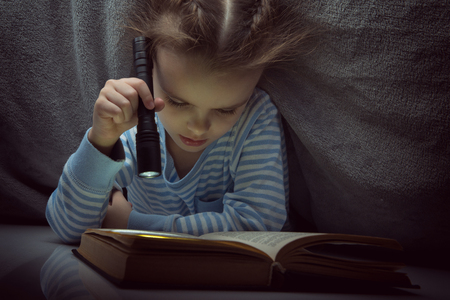Little girl reading fairy tales book under the covers at the evening with lantern. Cute kid playing before going to sleep, image toned.の写真素材