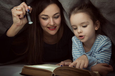 Little girl and her mother reading fairy tales book under the covers at the evening with lantern. Cute kid playing before going to sleep, image toned. Home family leisure.の写真素材