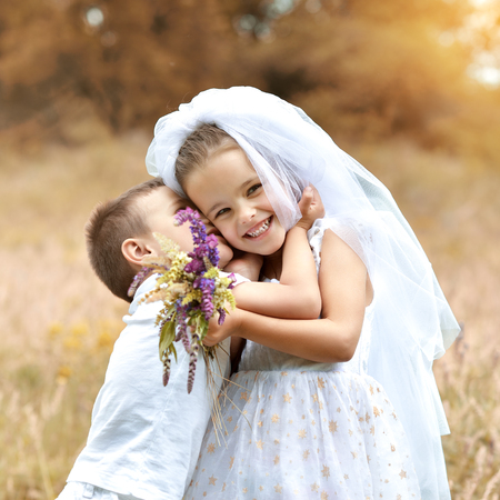 Young bride and groom playing wedding summer outdoor. Children like newlyweds. Little girl in bride whote dress and bridal veil kissing her little boy groom, kids game. Bridal, wedding concept.の写真素材