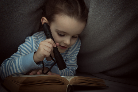 Little girl reading fairy tales book under the covers at the evening with lantern. Cute kid playing before going to sleep, image toned.の写真素材