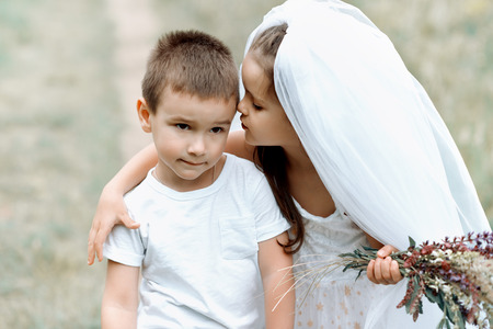 Young bride and groom playing wedding summer outdoor. Children like newlyweds. Little girl in bride white dress and bridal veil kissing her little boy groom, kids game. Bridal, wedding concept.の写真素材