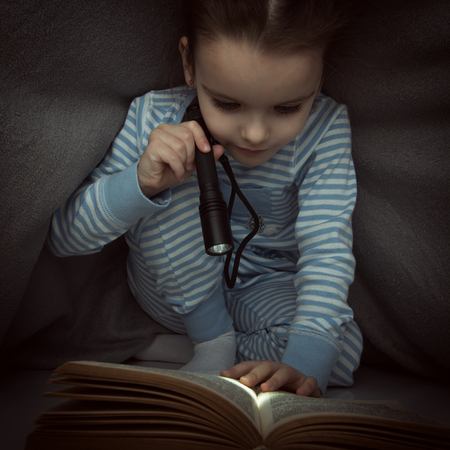 Little girl reading fairy tales book under the covers at the evening with lantern. Cute kid playing before going to sleep, image toned.の写真素材