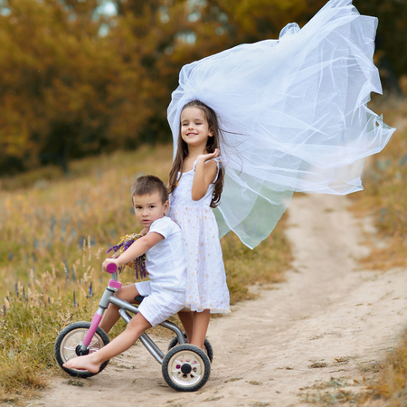 Young bride and groom playing wedding summer outdoor. Children like newlyweds on bicycle. Little girl in bride white dress and bridal veil walking with her little boy groom, kids game. Bridal, wedding concept, image toned and noise added.の写真素材