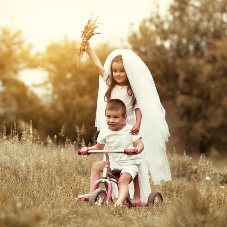 Young bride and groom playing wedding summer outdoor. Children like newlyweds on bicycle. Little girl in bride white dress and bridal veil walking with her little boy groom, kids game. Bridal, wedding concept, image toned and noise added.の写真素材