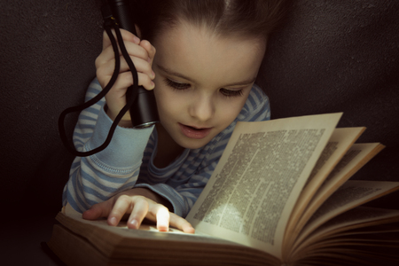 Little girl reading fairy tales book under the covers at the evening with lantern. Cute kid playing before going to sleep, image toned.の写真素材
