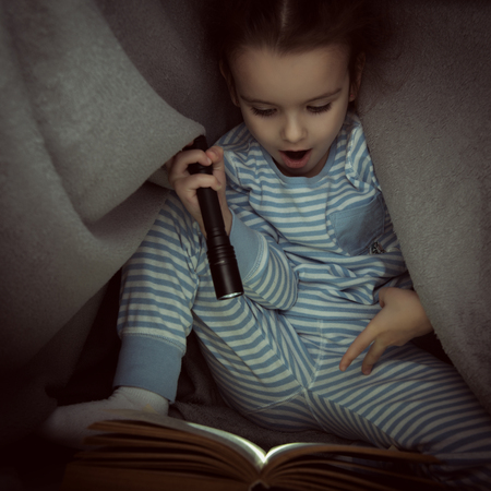Little girl reading fairy tales book under the covers at the evening with lantern. Cute kid playing before going to sleep, image toned.の写真素材