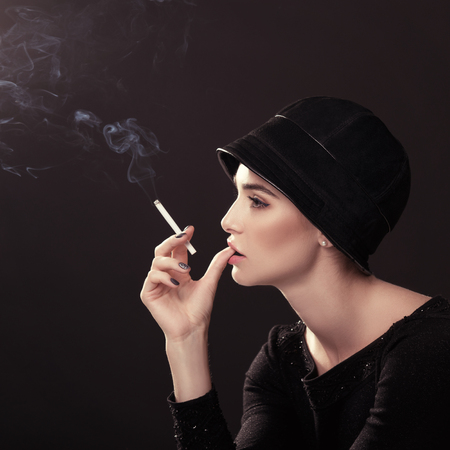 Young fashion woman smoking cigarette in hat and black drees over dark background. Vintage female...