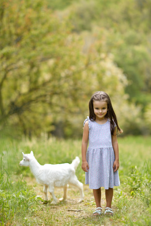 Little girl plays and huhs goatling in country, spring or summer nature outdoor. Cute kid with baby animal, countryside outdoor portrait, forest, trod, glade background. Friendship of child and yeanling, image toned.の写真素材