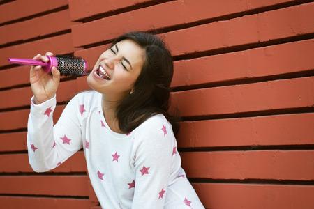 Young woman singing outdoor with comb for hair against red brick wall. Urban lifestyle, imagination, dreams, music, happiness, joy, teenage concept. Image toned and noise added.の写真素材