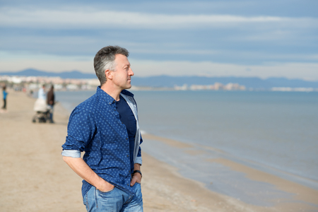 Handsome middle-aged man walking at the beach. Attractive mid adult male model posing at seaside in blue jeans, t-shirt shirt. Outdoor portrait of beautiful macho man.の写真素材