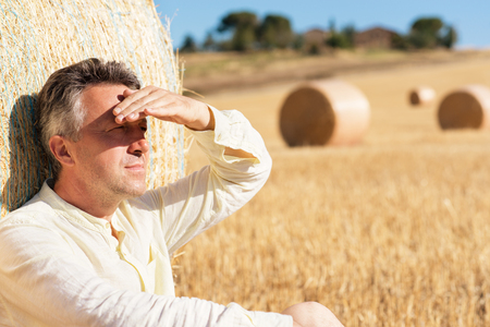Farmer sitting on field near bale of wheat. Harvestimg in Tuscany, Italy. Stacks of hay on summer field. Hay and straw bales in the end of summer. Working man resting in shadow after harvestingの写真素材