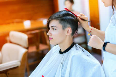 Beauty, hairstyle, treatment, hair care concept, young woman and hairdresser cutting hair at hairdressing salon. Hairdresser cutts beautiful happy girl's hair. Hairstylist serving client at barber shop. Professional barber doing hairstyle in barbershop, soft focus, image toned and noise added.の写真素材