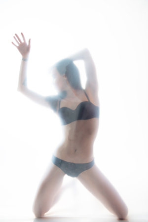 Young beautiful woman posing in lingerie behind translucent material. Full-length attractive sexy girl's portrait in studio over white background, foggy silhouette. Art female portrait.の写真素材
