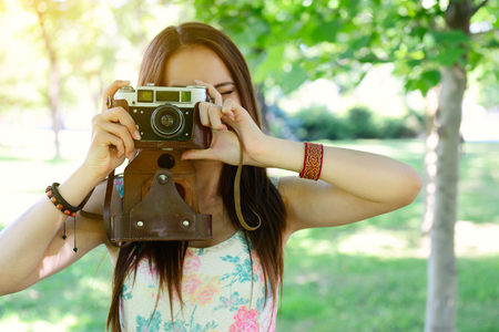 Cheerful beautiful girl talking pictures with vintage camera in summer park. Young woman photographer posing with her camera outdoor.の写真素材