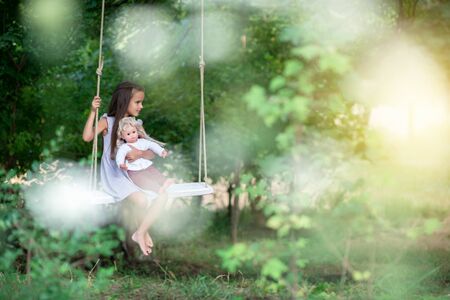 Happy girl rides on a rope swing with her retro doll in park. Little Princess has fun outdoor, summer nature outdoor. Childhood, child lifestyle, enjoyment, happiness.の写真素材
