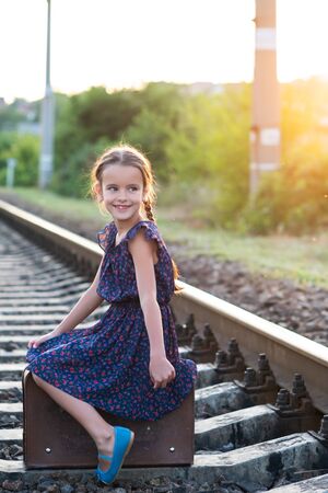 Beautiful charming little girl with pigtails dressed in dark blue dress with flowers and blouse sitting in big vintage luggage at railroad and happy smiling. Fashion, retro stylization.の写真素材