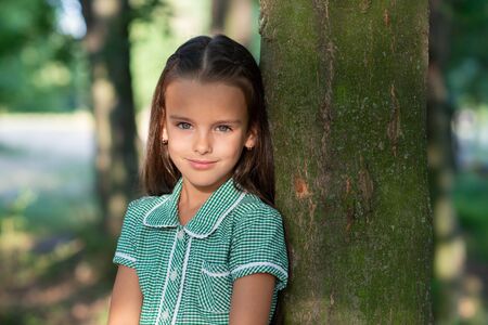 Pretty little brunette girl in forest near tree with sun light at her beautiful face. Childhood. Cute kid outdoor portrait.の写真素材