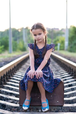Beautiful charming little girl with pigtails dressed in dark blue dress with flowers and blouse sitting in big vintage luggage at railroad. Fashion, retro stylization.の写真素材