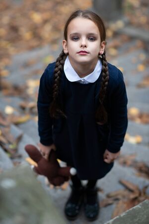 Beautiful sad little girl with with pigtails, dressed in dark blue standing near mystic abandoned building with gothic stairs and holding handmade bear toy. Halloween horror, ghost or spirit of child. Loneliness, depressionの写真素材