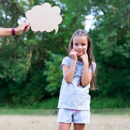 Pretty thinking little girl puts on a happy face, summer nature outdoor with cloud of thoughts (like in comic book). Kid's portrait. Emotions, feeling and thoughts of childの写真素材