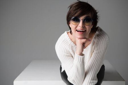 Young woman with sunglasses sitting on white cube in studio isolated over gray, looking at camera...