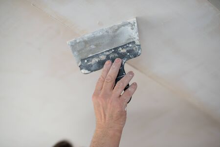 Putty knife in male hand. Hand workÑ Worker putsty plasterboard ceiling in new appartment. Repairman works with plasterboard, plastering dry-stone wall, home improvement. Man makes repairs at house.の写真素材