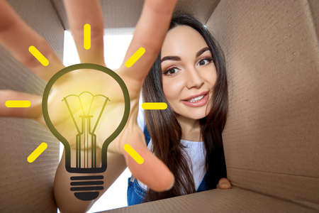 Happy girl opening postal package with light idea cartoon lamp inside. Excited young woman finds an idea bulb in cardboard box. Gift, present, delivery, shipment, sale, brilliant solution.の写真素材