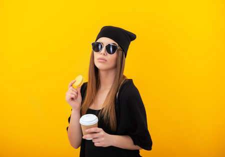 Stylish girl eats cake macaron over yellow background. Studio female portrait with sweet food and drink ,take and go. Woman drinks coffee and eats dessertの写真素材