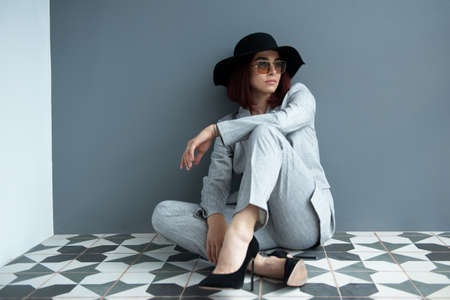 Beautiful fashion woman. Full length portrait of beautiful girl wearing gray suit and black hat with sun glasses sitiing on the floor at homeの写真素材